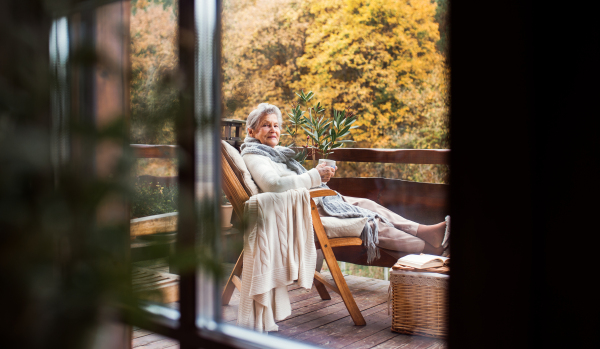 An elderly senior woman sitting outdoors on a terrace in on a sunny day in autumn. Shot through glass.