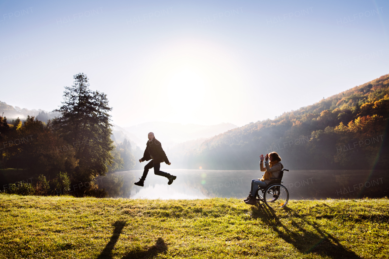 Active senior couple on a walk in a beautiful autumn nature. A woman in a wheelchair and a man jumping by the lake in the early morning.