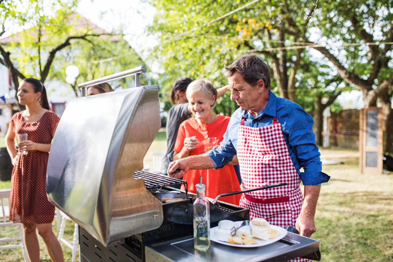 Family celebration outside in the backyard. Barbecue party. An old man cooking food on the grill.