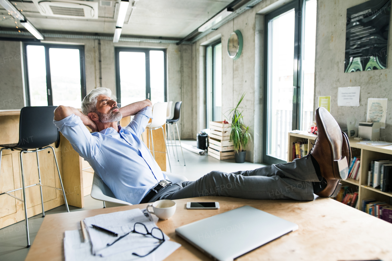 Thoughtful mature businessman with gray hair in the office with feet up on desk. Man with smartphone and laptop on the desk.