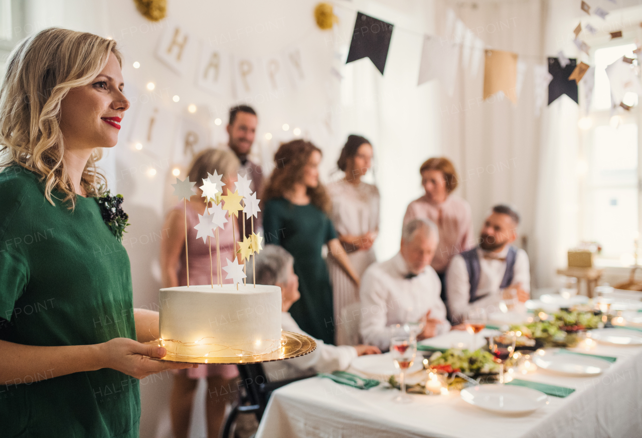 A young woman holding a birthday cake on an indoor party, multigeneration family in the background.