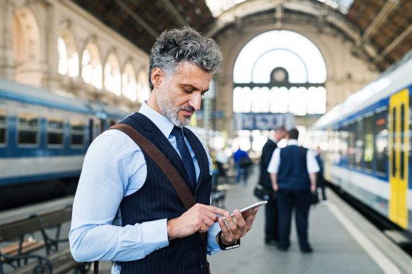 Handsome mature businessman with smartphone in a city. Man waiting for the train at the railway station, text messaging.