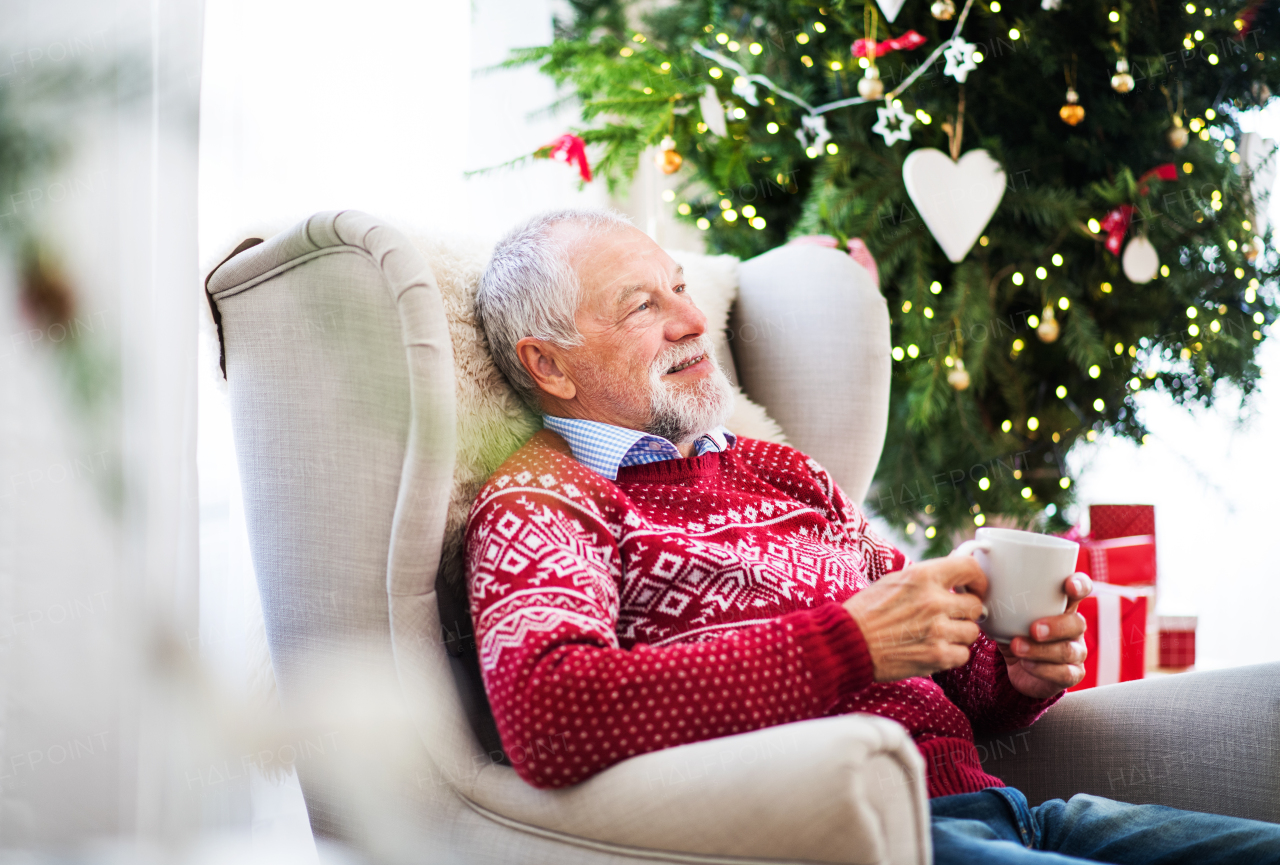 A portrait of senior man sitting on armchair at home at Christmas time, holding a white cup.