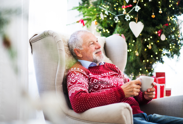 A portrait of senior man sitting on armchair at home at Christmas time, holding a white cup.