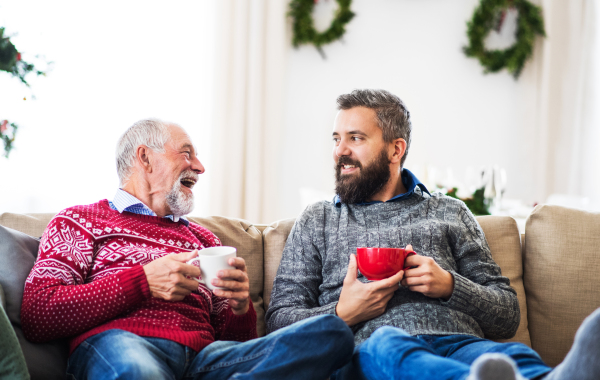 A senior father and adult son with feet on a coffee table sitting on a sofa at home at Christmas time, holding a cup of coffee and talking.