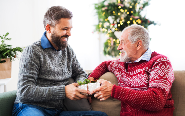 A senior father and adult son sitting on a sofa at home at Christmas time, giving presents to each other.
