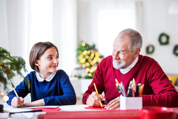 A small girl and her grandfather sitting at a table, writing Christmas cards together.