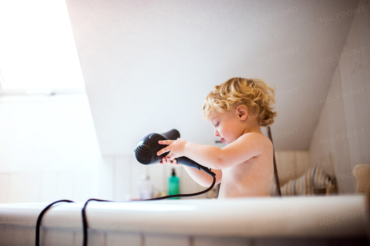 Cute little toddler boy with a hairdryer standing in the tub in the bathroom. Dangerous situation. Domestic accident.