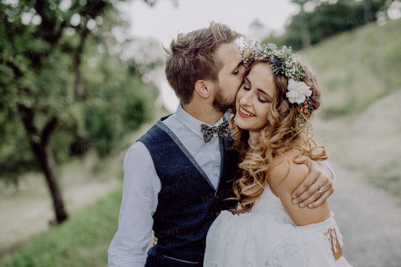 Beautiful young couple, groom and bride with flower wreath outside in green nature. A man kissing a woman.
