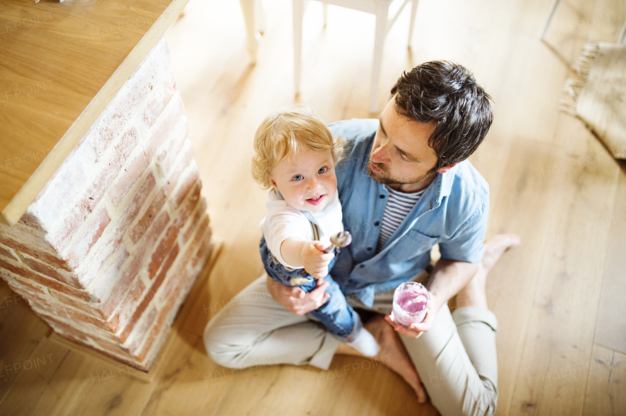 Young father at home sitting on the floor feeding his cute little son with yoghurt.