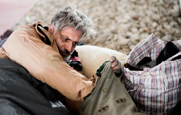 A homeless beggar man lying on a ground outdoors in city, taking something out of a bag.