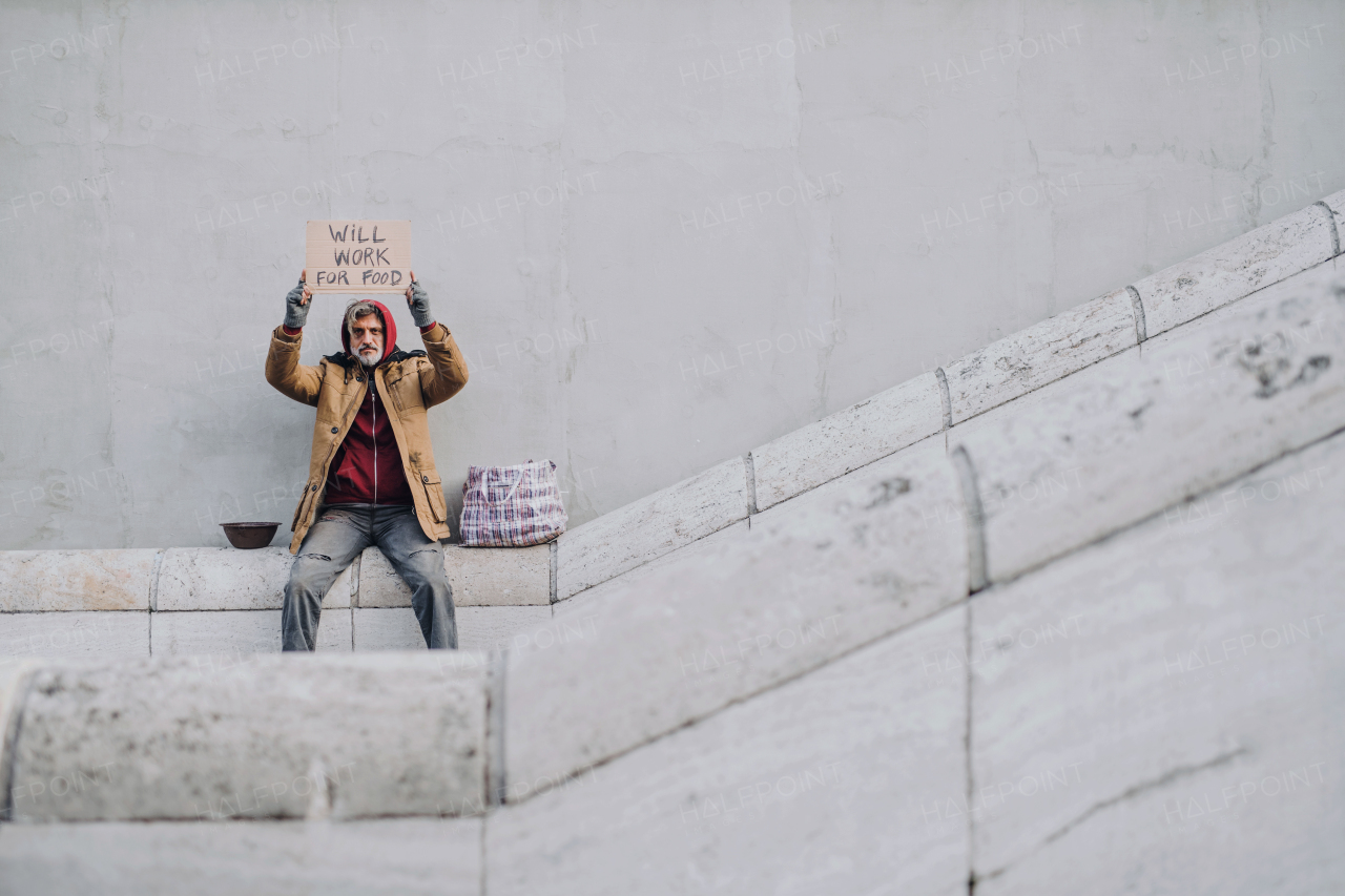 Homeless beggar man sitting outdoors in city holding will work for food cardboard sign, asking for money donation. Copy space.