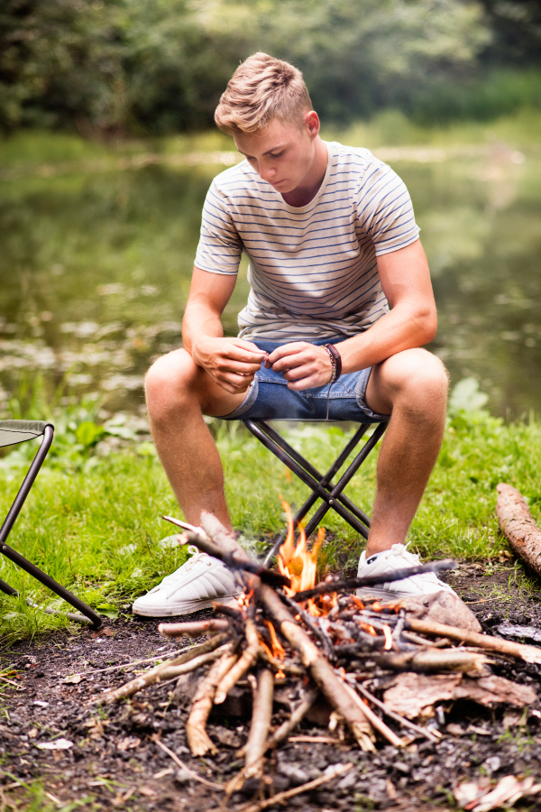 Teenage boy enjoying camping vacations in forest, sitting at bonfire.