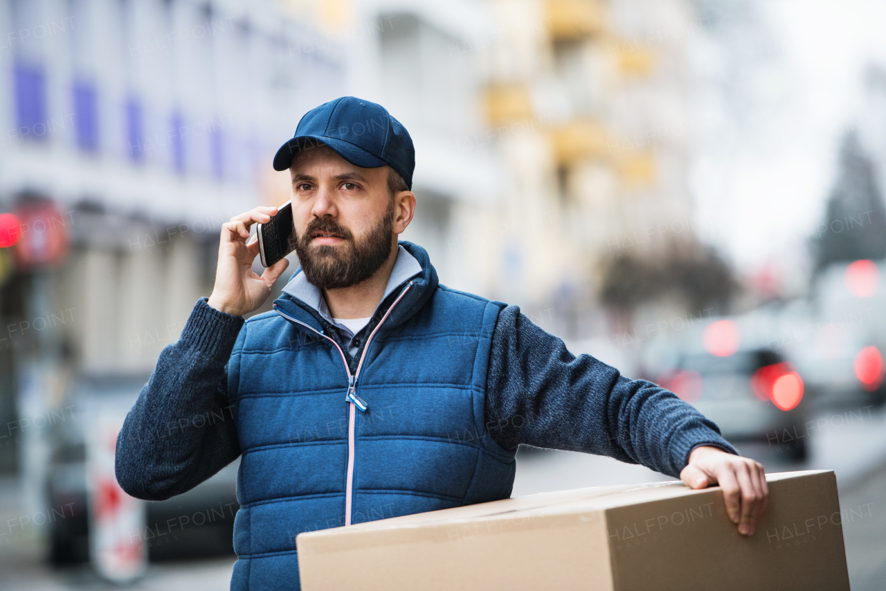 Delivery man holding a parcel box on the street - courier service concept. A man with a smartphone making a phone call.