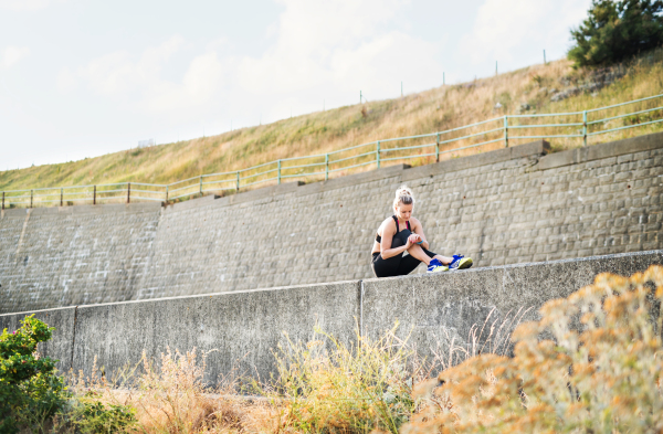 Young sporty woman runner sitting on a concrete wall outside, using smartwatch and resting.
