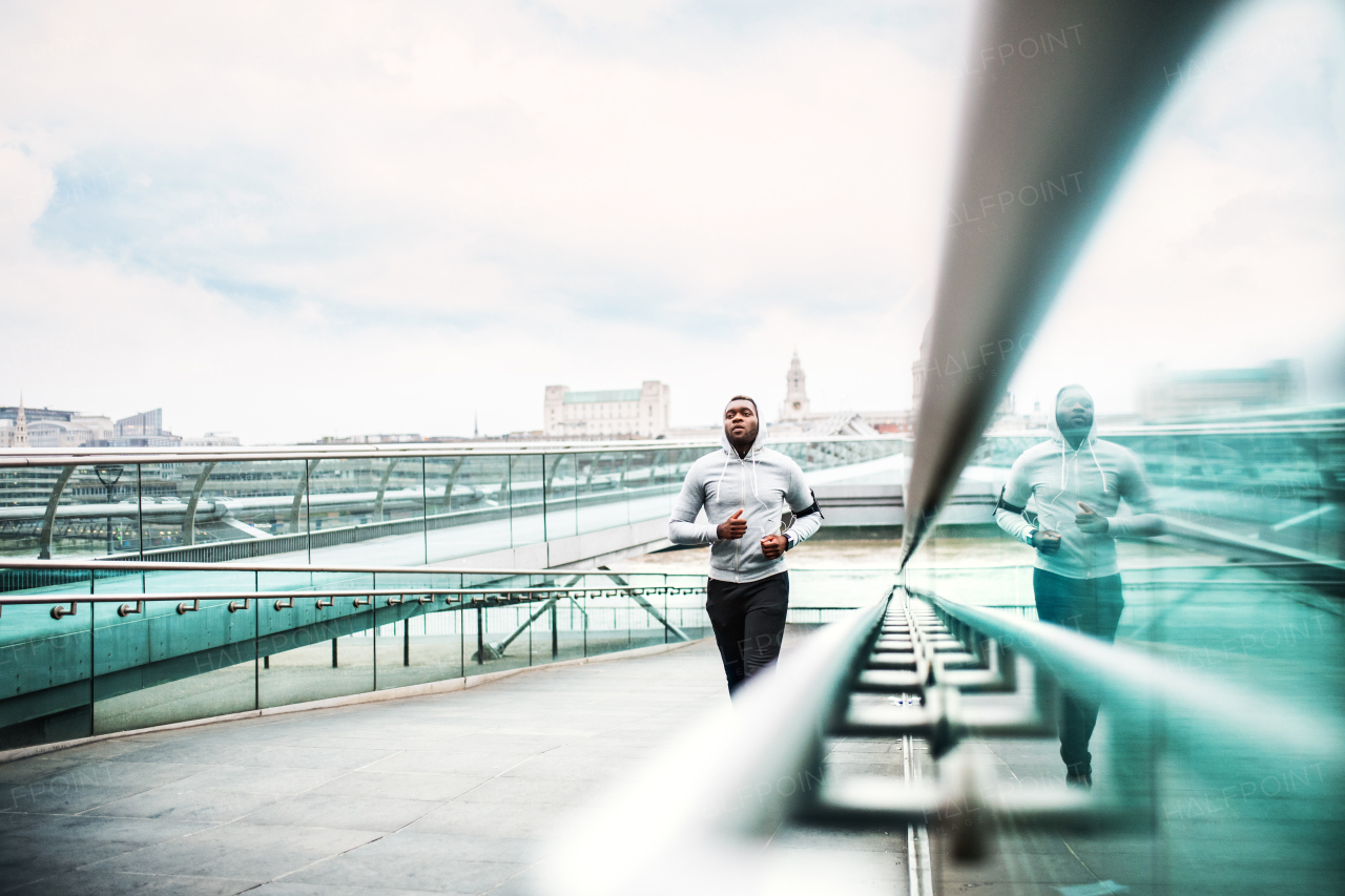 Young sporty black man runner with smartphone in armband and earphones running on the bridge outside in a city. Copy space.