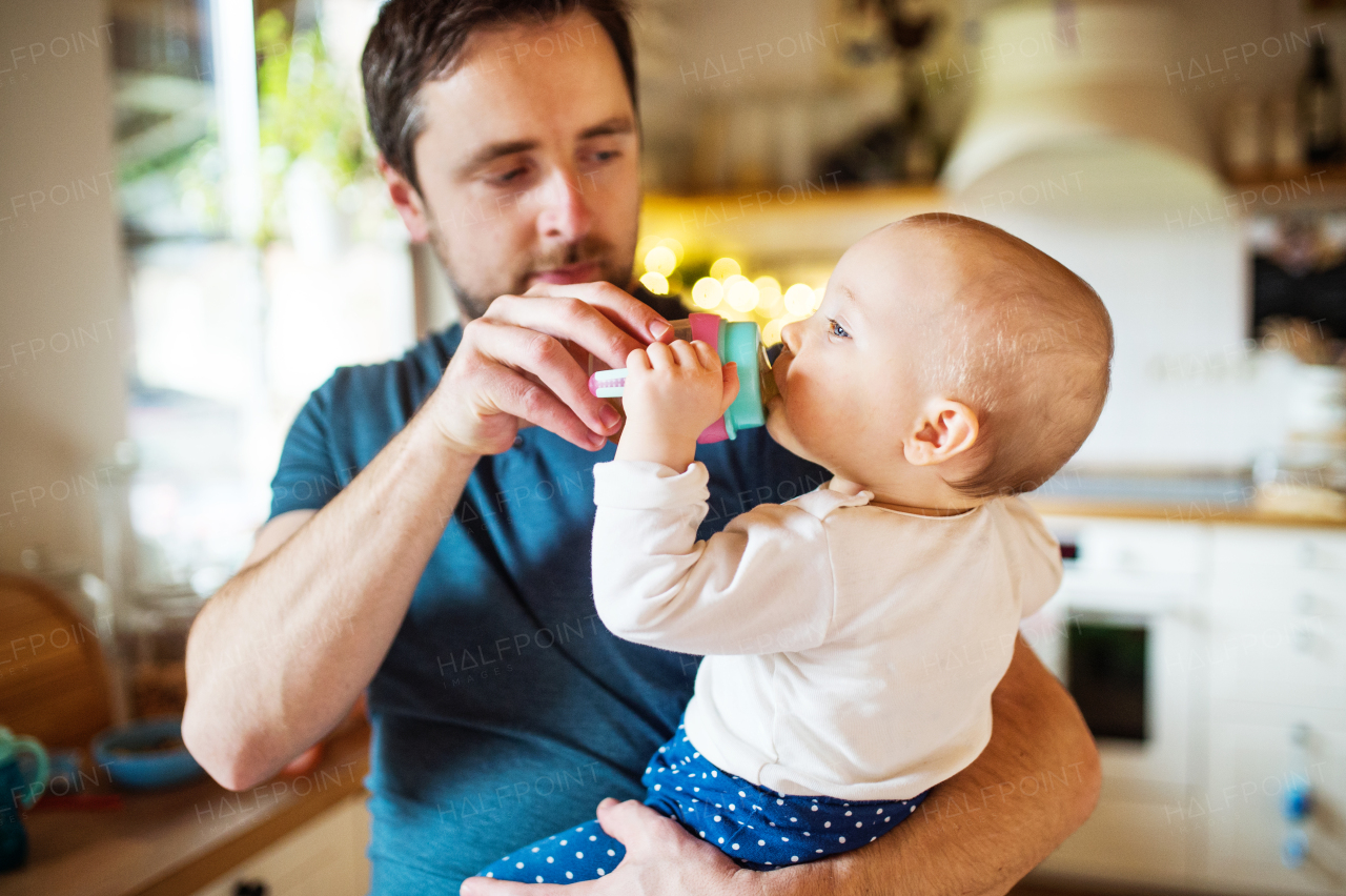A father with a baby daughter at home. A cute girl drinking water from the bottle. Paternity leave.