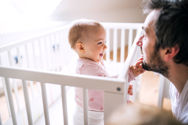 A small toddler girl standing in cot touching her fathers face at home. Paternity leave.