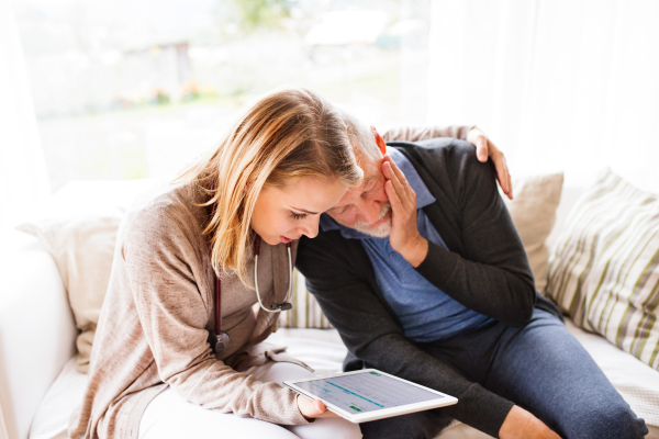 Health visitor and a senior man during home visit. A female nurse or a doctor showing test results on a tablet.
