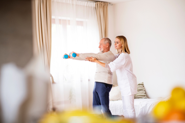 Health visitor and a senior man during home visit. A nurse or a physiotherapist helping a senior man exercise with dumbbells.