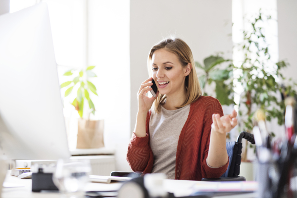 Businesswoman in wheelchair with smartphone sitting at the desk, making a phone call.
