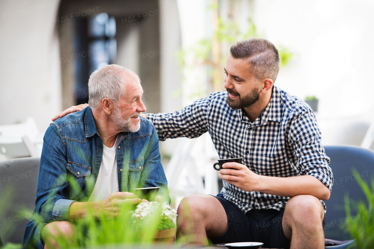 An adult hipster son and senior father with a cup of coffee in an outdoor cafe, talking.