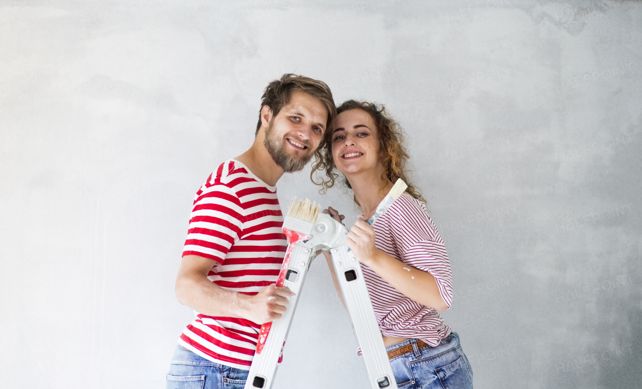 Beautiful young couple standing on ladder painting walls in their new house using paint brushes. Home makeover and renovation concept.