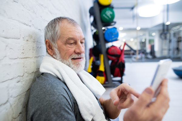 Fit senior man in sports clothing in gym resting after working out, towel around his neck, holding smart phone, texting