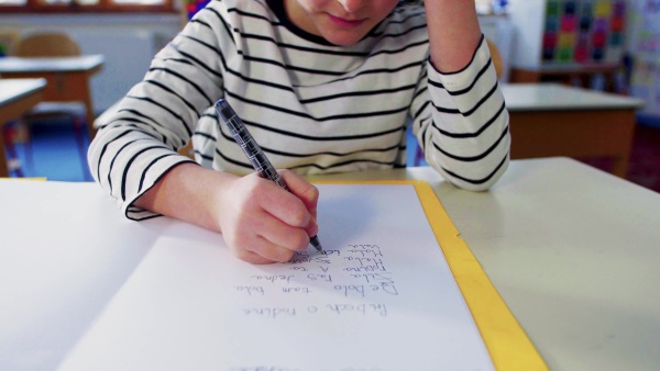 An unrecognizable small girl sitting at the desk at school writing.