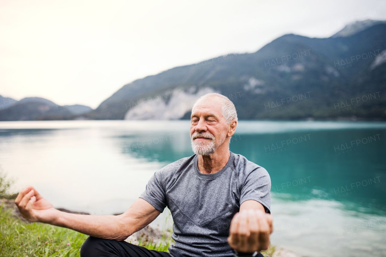A senior man pensioner sitting by lake in nature, doing yoga exercise. Copy space.