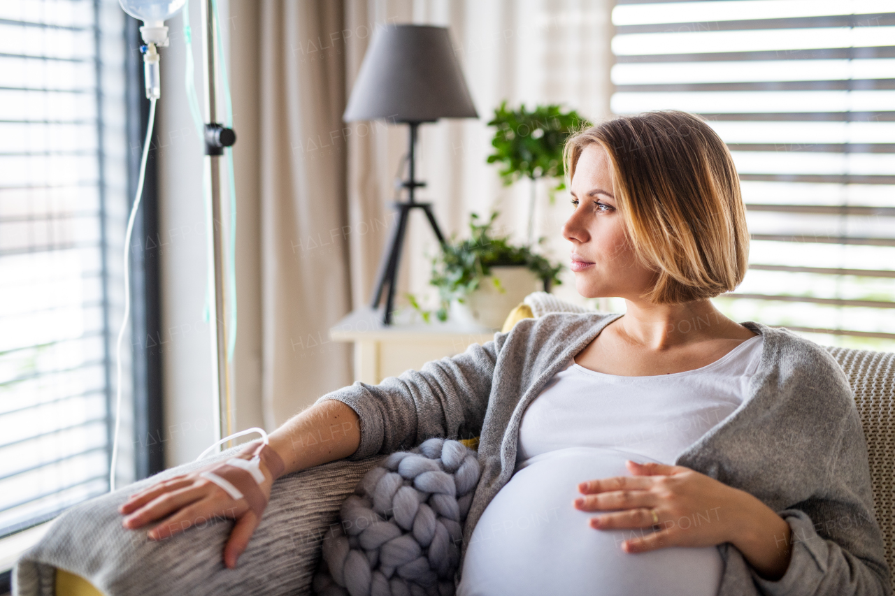 A portrait of pregnant woman with IV drip indoors at home or in hospital, sitting on sofa.