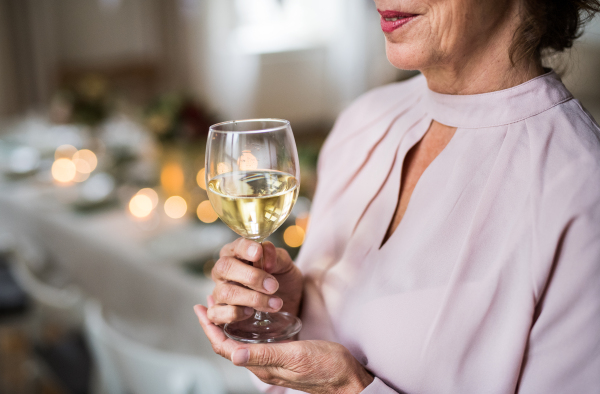 A midsection of a senior woman standing indoors in a room set for a party, holding a glass of wine. Copy space.