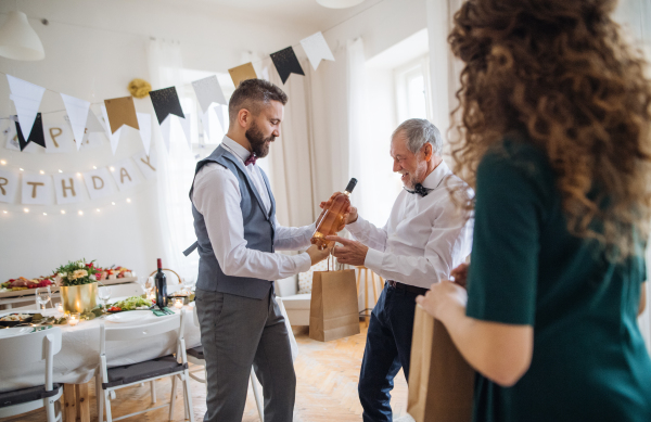 A mature man giving a bottle of wine to his father on indoor birthday party, a celebration concept.