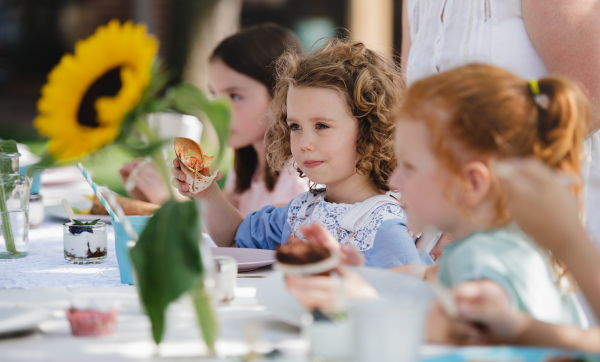 Happy small girls sitting and eating at table on summer garden party, birthday celebration concept.