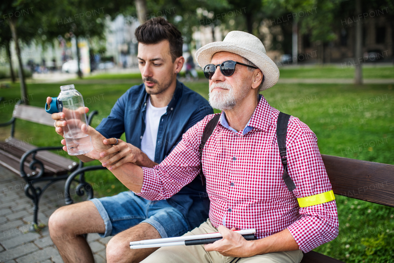 A young man and blind senior with white cane and water bottle sitting on bench in park in city.