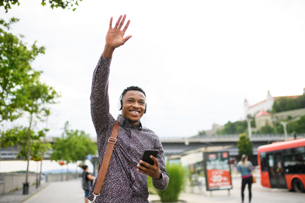 Cheerful young black man commuter with smartphone outdoors in city, greeting somebody.