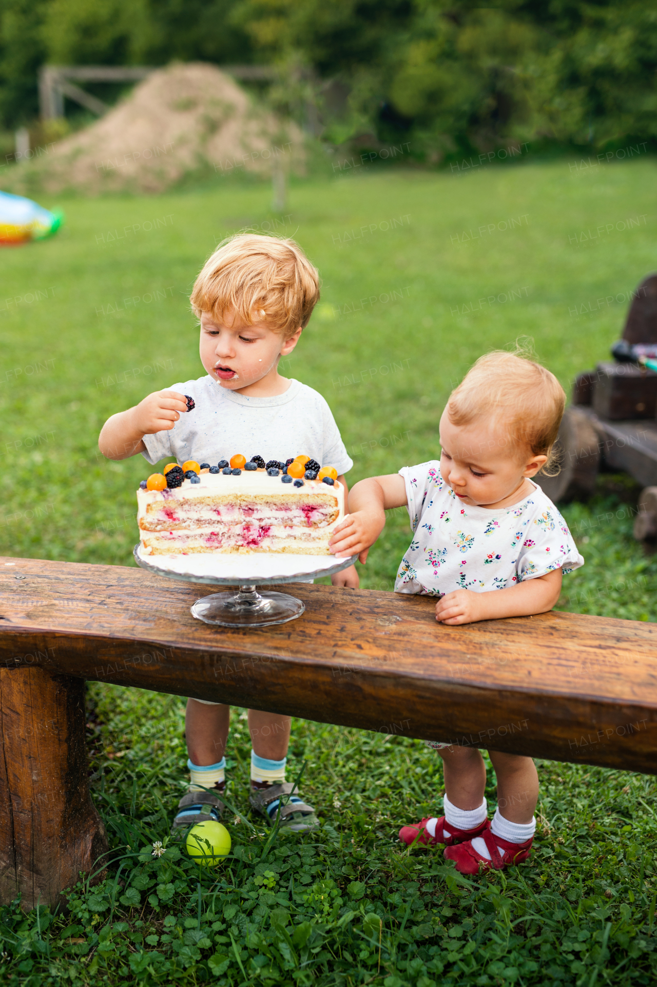 Front view of two toddler children with birthday cake outdoors in garden in summer.
