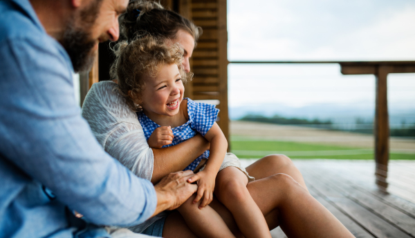 Happy family with small daughter sitting on patio of wooden cabin, holiday in nature concept.
