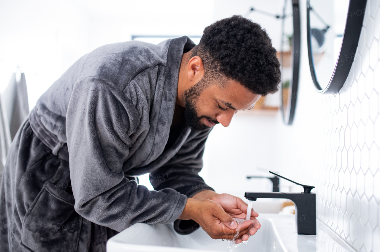 Happy young man washing face ndoors at home, morning or evening routine concept.