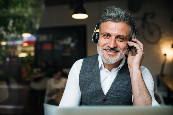 Mature man with headphones at the table in a cafe, using laptop. Shot through glass.