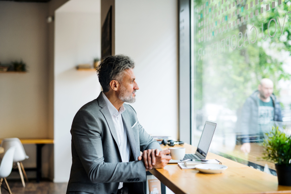 Mature man with coffee and laptop at the table in a cafe, looking out of window.