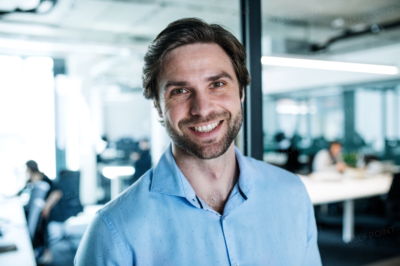 A portrait of young businessman standing in an office, looking at camera.