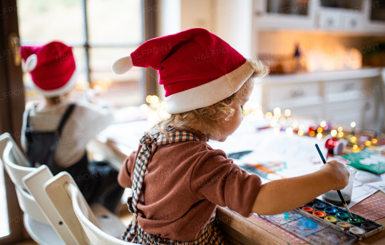 Rear view of happy small girl and boy indoors at home at Christmas, painting pictures.