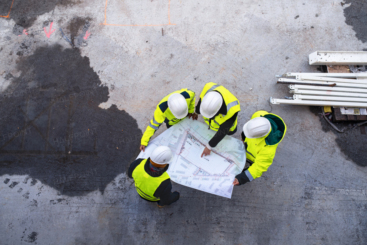 A top view of group of unrecognizable engineers with blueprints standing on construction site.
