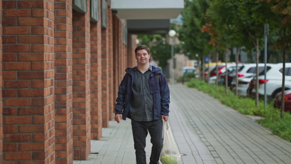 A man with Down syndrome walking outdoors in town street, holding mesh shopping bag and looking at camera.