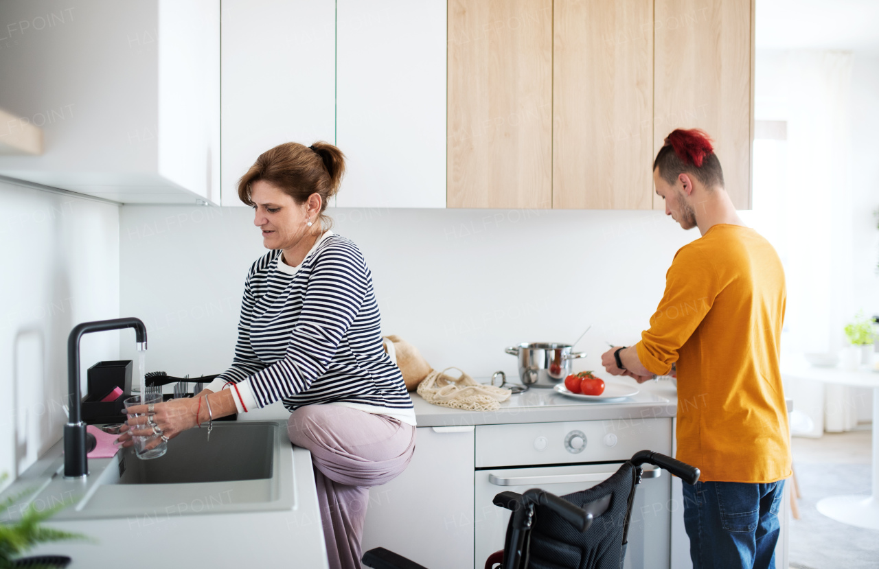 A disabled mature woman in wheelchair talking to son in kitchen indoors at home.