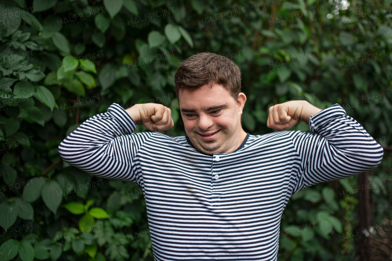 Portrait of down syndrome adult man standing outdoors at green leaves background, flexing muscles.