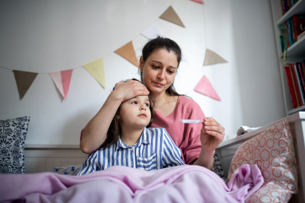 Portrait of mother looking after sick small daughter in bed at home, measuring temperature.