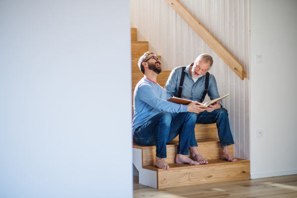 An adult hipster son and senior father sitting on stairs indoors at home, looking at photographs.