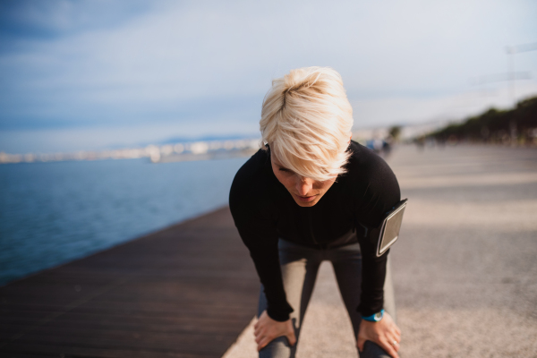 Front view portrait of young sportswoman with smatphone standing outdoors on beach, resting.